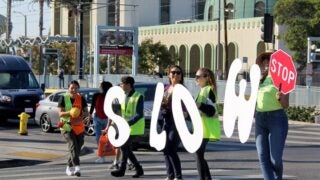 Students holding cut out letters that spell the word 'slow' while helping kids cross the street.