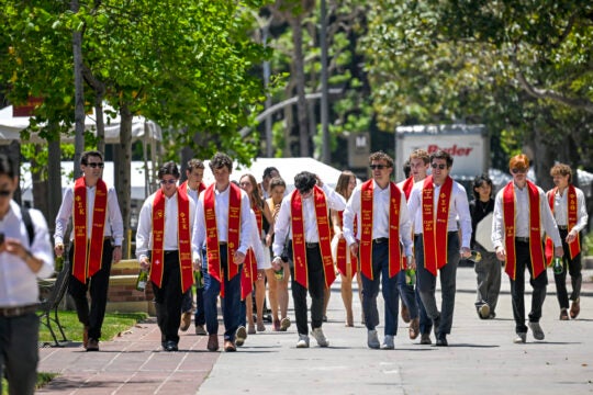 A group of men wearing uniform outfits of white shirts, slacks, and their graduation sashes walk down Trousdale parkway.