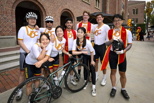 A group of Trojans wearing their biking outfits pos in front of Bovard Building with their graduation sashes