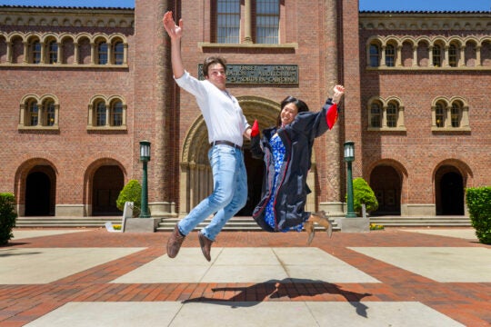 USC Thornton grads Heeseung Lee, right, and Victor Martinez Sara jump for a photograph in front of Bovard